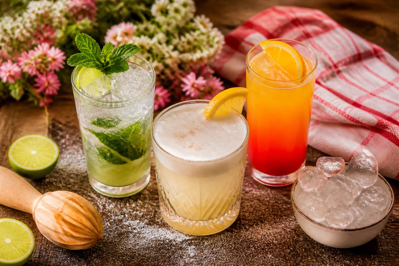Drei fertige Cocktails auf einem Holztisch – ein Mojito mit Minze, ein Sour mit Schaumhaube und ein Tequila Sunrise mit Farbverlauf, daneben halbierte Limetten und eine Schale Eis (mit AI erstelltes Bild)