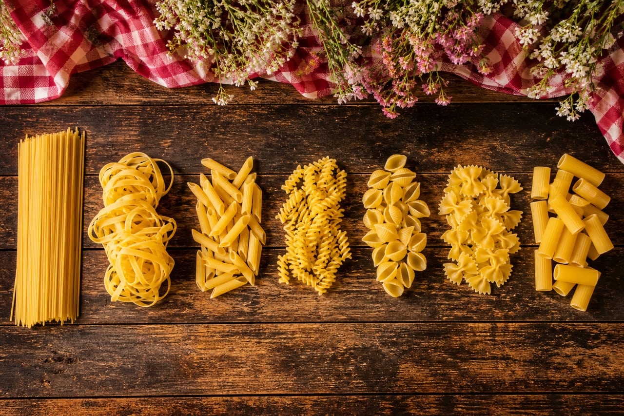 Sieben italienische Nudelsorten in getrennten Häufchen: Spaghetti, Tagliatelle, Penne, Fusilli, Conchiglie, Farfalle und Rigatoni (mit AI erstelltes Bild)